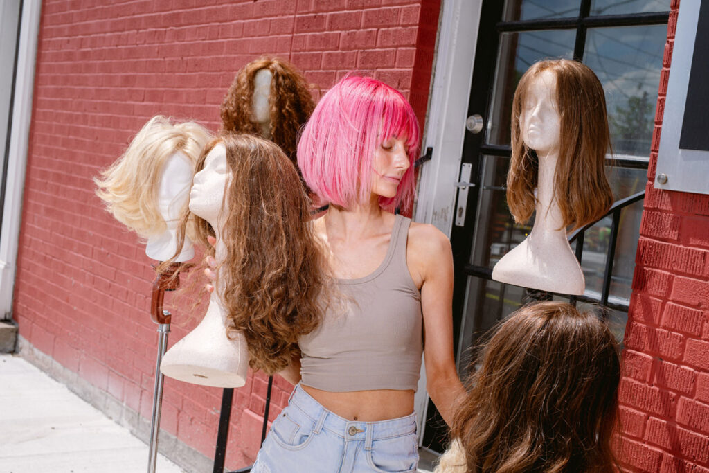 Woman wearing pink wig holding and surrounded by mannequins wearing wigs